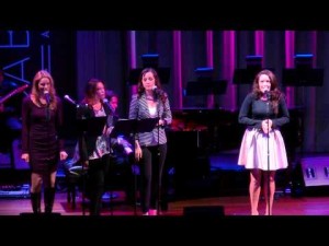 Three women singing on stage with a pianist in the background.
